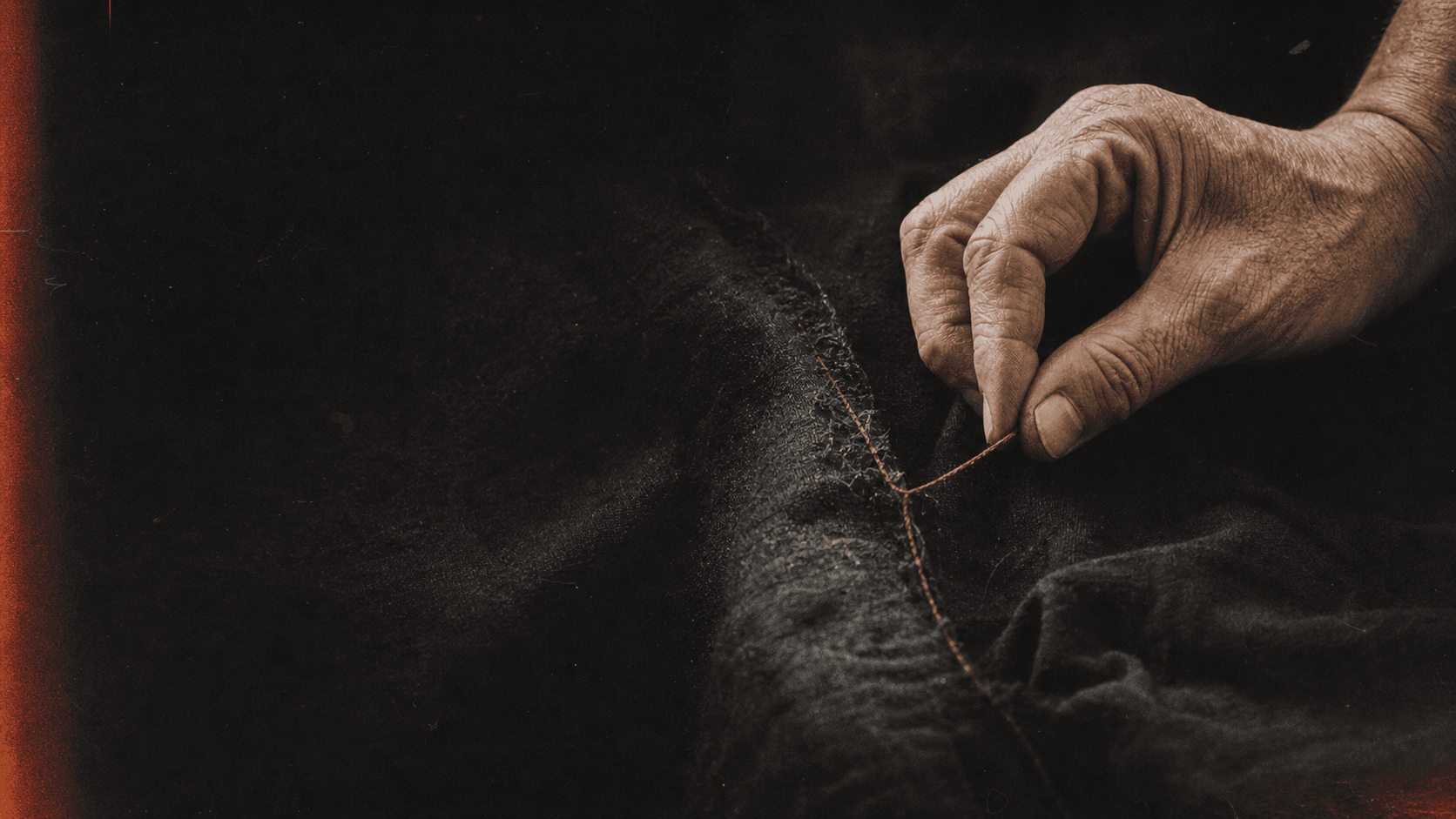 A weathered hand drawing red thread through dark cloth